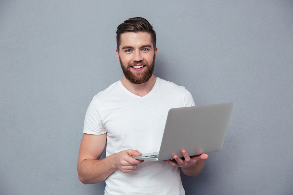 Portrait of a smiling casual man holding laptop over gray background-1